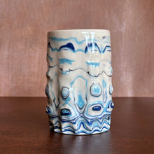 Load image into Gallery viewer, A handmade ceramic vase with a textured, wavy surface featuring a blue and white marbled pattern, set on a dark wooden table against a warm brown background.