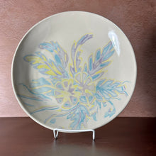 Load image into Gallery viewer, A round shallow ceramic bowl, decorated with a colorful abstract floral pattern in shades of blue, yellow, and purple. The plate is displayed on a small white metal stand against a textured brown wall and dark brown tabletop.