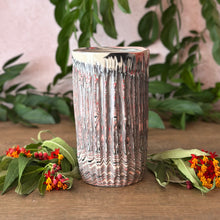 Load image into Gallery viewer, A cylindrical ceramic vase with a textured, vertically ribbed surface featuring a marbled pattern of black, white, and white. It is styled on a wooden surface with green leaves and small red and orange flowers against a soft pink background.