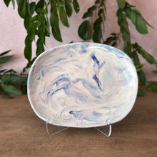 Load image into Gallery viewer, An oval ceramic plate with a white, blue, and purple marbled pattern, displayed on a clear plastic stand on a wooden surface with green leaves in the background.