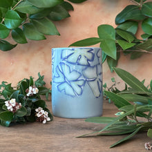 Load image into Gallery viewer, Blue ceramic cup with floral design on a wooden surface surrounded by green leaves