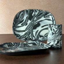 Load image into Gallery viewer, A set of black and white marbled ceramic tableware, including a rectangular platter, a square bowl, and a cylindrical cup, displayed on a dark wooden table against a textured beige wall.
