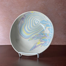 Load image into Gallery viewer, A shallow ceramic bowl with a swirling marbled pattern in light blue, lavender, and yellow on an off-white base, displayed on a small white stand against a textured pink wall and dark brown surface.
