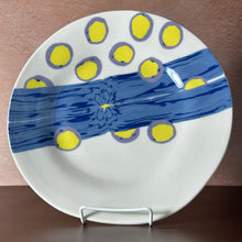 Load image into Gallery viewer, A white ceramic plate with a blue river design and yellow circles outlined in purple, displayed on a small metal stand against a brown background.
