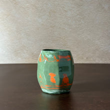 Load image into Gallery viewer, A small green ceramic vase with orange hand-painted abstract designs, sitting on a dark wooden table against a textured beige wall.
