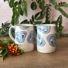 Load image into Gallery viewer, Two ceramic mugs with a blue, purple, and white abstract swirl pattern on a white base, displayed on a wooden table with green leaves and small red flowers.

