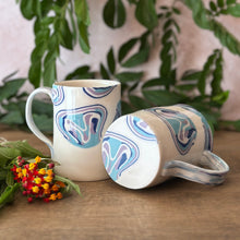 Load image into Gallery viewer, Two ceramic mugs with a blue, purple, and white abstract swirl pattern, displayed on a wooden surface with green leaves and small flowers in the background.
