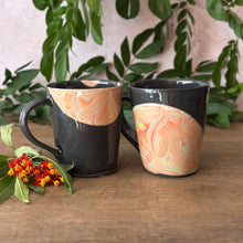 Load image into Gallery viewer, Two ceramic mugs with a black exterior and peach, cream, and orange marbled pattern on surface,  displayed on a wooden surface with green leaves and small flowers.
