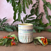 Load image into Gallery viewer, A handmade ceramic cup with a marbled pattern of light blue, cream, and yellow, styled on a wooden surface with green leaves and small red and orange flowers against a soft pink background.
