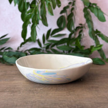 Load image into Gallery viewer, A shallow cream-colored ceramic bowl with a marbled pattern of light blue, yellow, and lavender swirls on the exterior, resting on a wooden surface with green foliage and a pink wall in the background.
