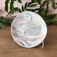 Load image into Gallery viewer, A round ceramic plate with a marbled swirl pattern of blue, purple, and white , displayed on a clear stand on a wooden surface with green leaves in the background.
