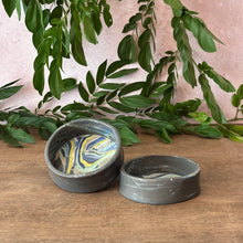 Load image into Gallery viewer, Two small oval ceramic dishes with a dark gray exterior and a marbled interior in blue, yellow, and white, displayed on a wooden surface with green leaves and a pink wall in the background.
