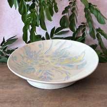 Load image into Gallery viewer, A ceramic bowl with a colorful floral pattern in blue, yellow, and purple on a cream background, displayed on a wooden surface with green plants in the background.
