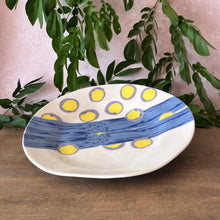 Load image into Gallery viewer, A ceramic serving bowl with a blue and yellow pattern, displayed on a wooden surface with green foliage and a pink wall in the background.
