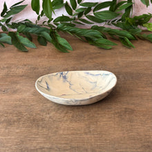 Load image into Gallery viewer, A small oval ceramic dish with a cream-colored glaze and blue marble veining, set on a wooden tabletop with green leaves in the background.
