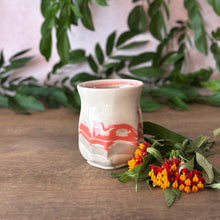 Load image into Gallery viewer, A small ceramic vase that is cream-colored  with a coral-pink abstract swirl pattern, styled on a wooden tabletop with green leaves and small red and yellow flowers.
