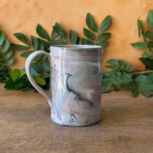 Load image into Gallery viewer, A ceramic mug with a marbled gray, white, and subtle pink pattern, sitting on a wooden table with green leaves and a textured orange wall in the background.
