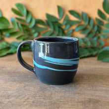 Load image into Gallery viewer, A handmade ceramic mug with a dark charcoal black base and light blue swirl patterns, resting on a rustic wooden surface with green leaves and a warm orange background.
