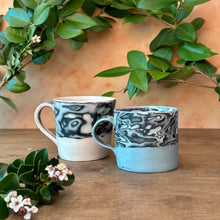 Load image into Gallery viewer, Two marbled ceramic mugs on a wooden surface with greenery in the background
