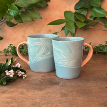 Load image into Gallery viewer, Two light blue ceramic mugs with orange handles on a wooden surface with greenery in the background.
