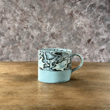 Load image into Gallery viewer, Ceramic mug with marbled design on a wooden surface and stone wall background
