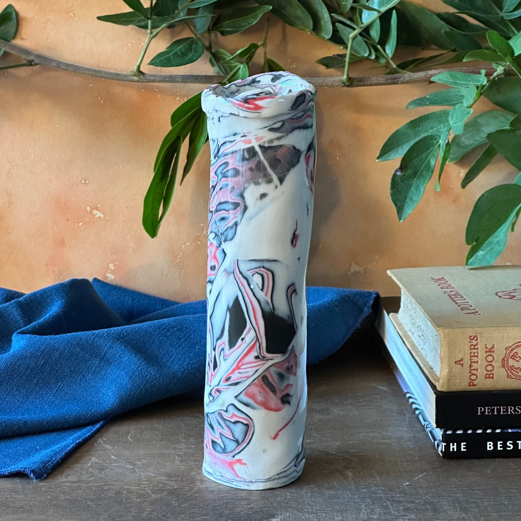 Tall cylindrical bud vase with abstract design on a wooden surface with books and a blue cloth.