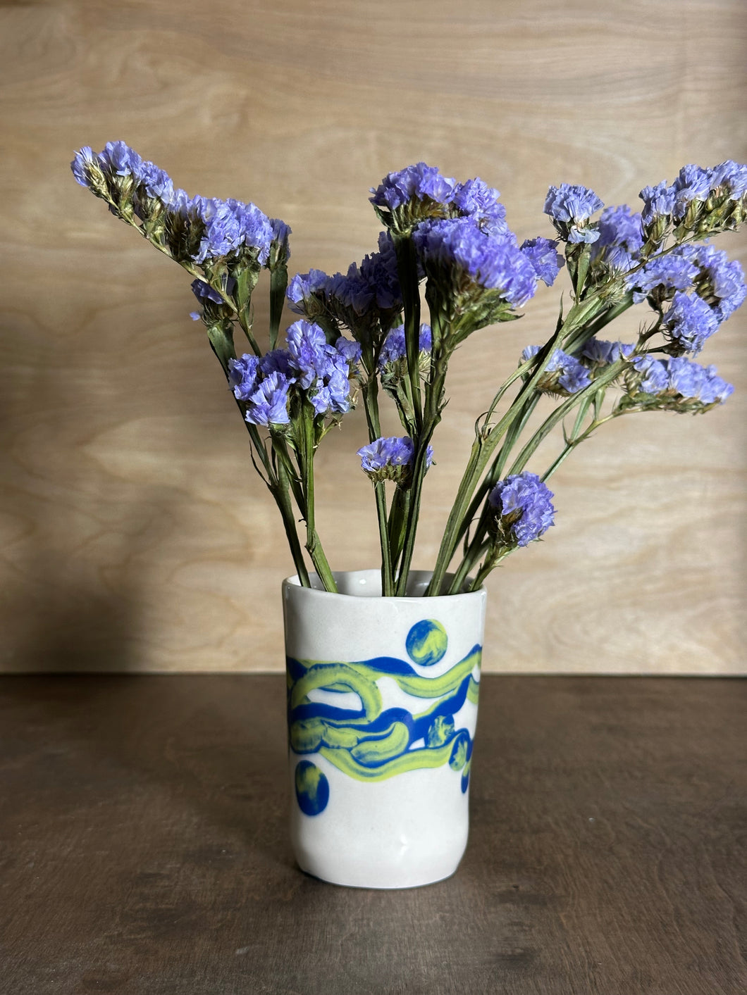 small white vase with blue and chartreuse flowing pattern holding purple flowers
