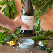 Load image into Gallery viewer, Bottle of Dierberg Pinot Noir wine held by a hand over a ceramic wine bottle coaster with greenery background
